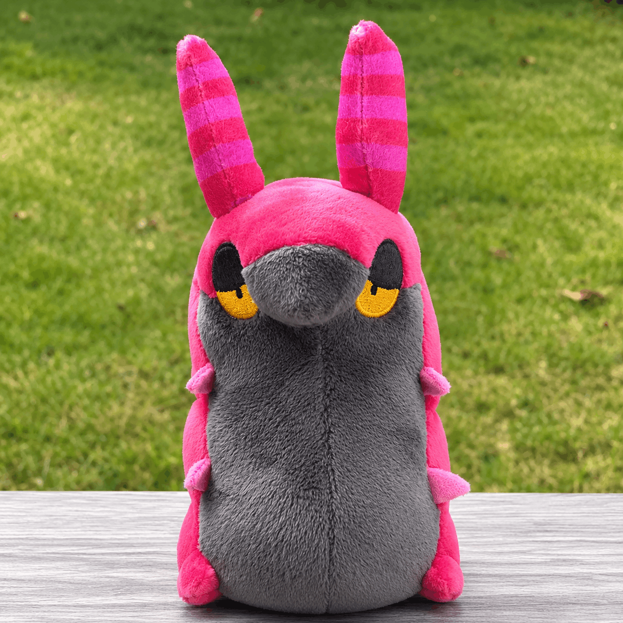 Pokemon Scolipede Plush Toy – Soft Centipede Stuffed Animal A pink and gray plush toy with long striped ears and yellow eyes, photographed sitting on a gray wooden surface with a blurred green grass background.