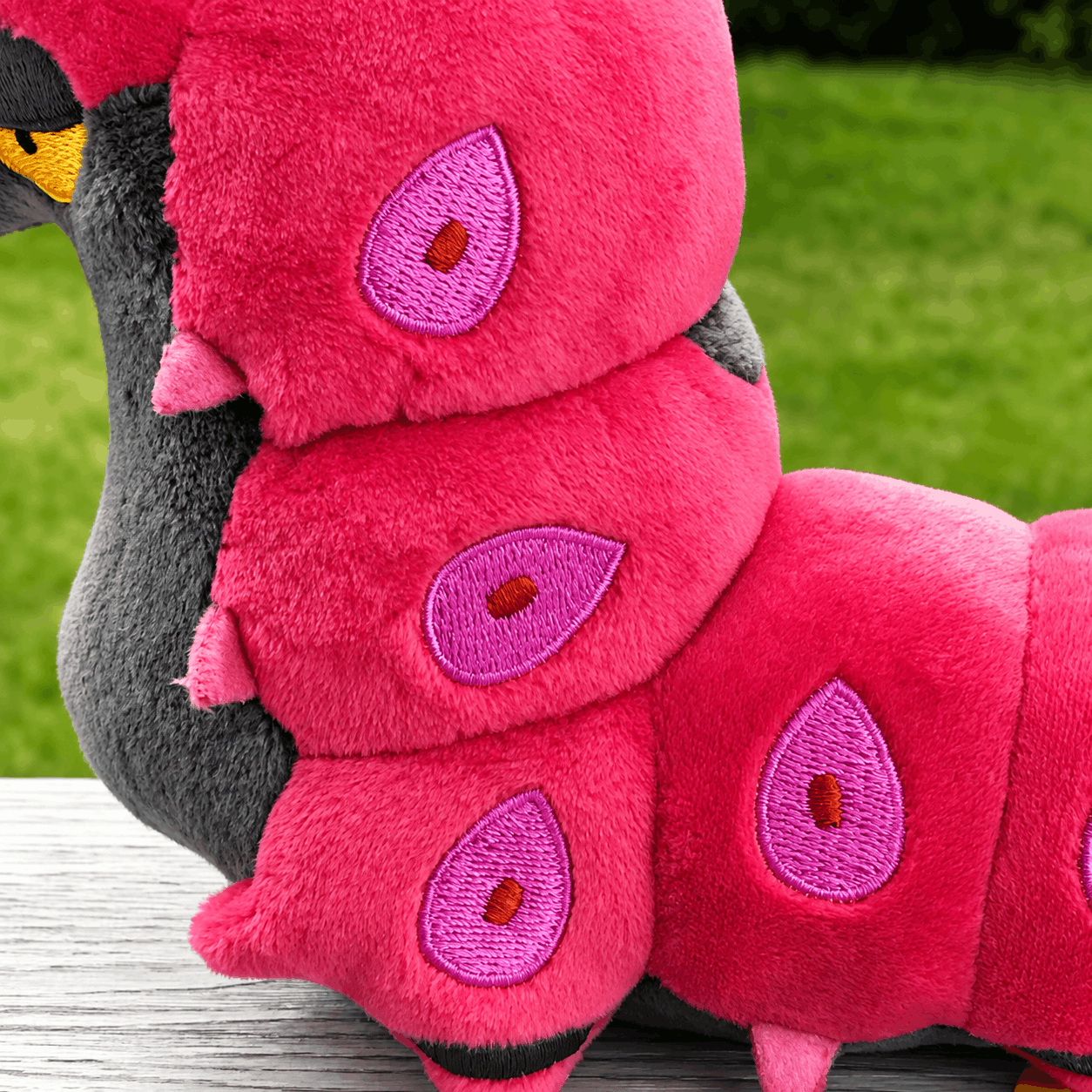 Pokemon Scolipede Plush Toy – Soft Centipede Stuffed Animal A close-up of a pink plush toy resembling a caterpillar or dragon, with embroidered purple teardrop motifs and a dark gray body, resting on a wooden surface with a blurred green grass background.