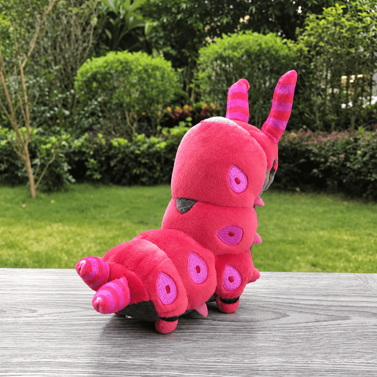 Pokemon Scolipede Plush Toy – Soft Centipede Stuffed Animal A pink caterpillar plush toy sitting on a gray wooden table with a green garden background.