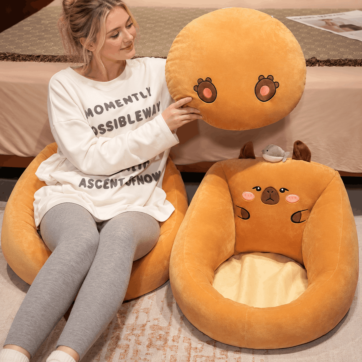 Capybara Floor Cushion – Futon Seat for Living Room & Bedroom A woman sits on a large round orange plush chair next to a matching shaped pet bed with a cute animal face design. The scene is indoors on a patterned rug in front of a bed.