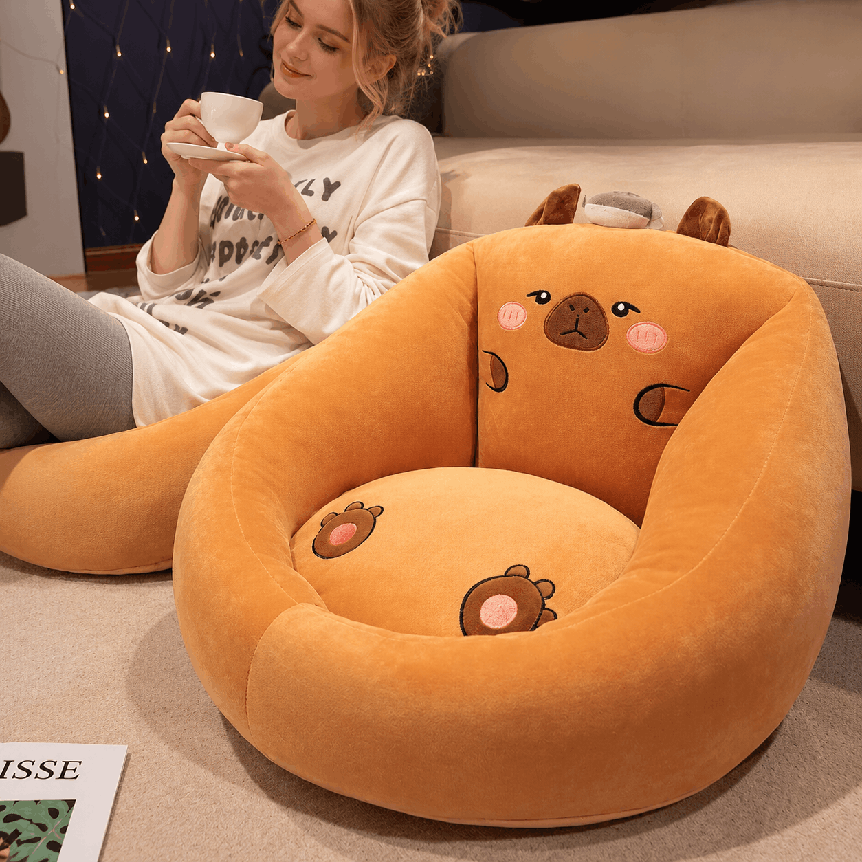 Capybara Floor Cushion – Futon Seat for Living Room & Bedroom A cozy orange plush capybara-shaped armchair in a living room setting, with a woman sitting in the background holding a cup and saucer.