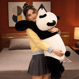 A woman in a yellow and gray sweatshirt and pleated skirt hugging a very large plush panda toy in a bedroom setting.