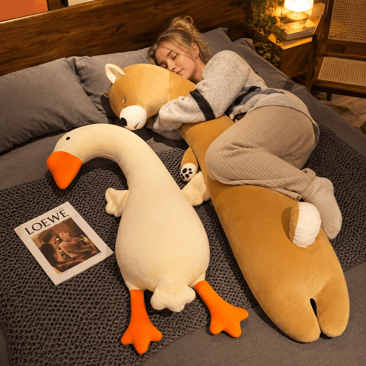 Squishmallow Fluffy Animal Body Pillow A cozy bedroom scene with a woman sleeping on a bed surrounded by plush toys, including a large tan animal-shaped pillow, a white goose plush with orange feet, and a fox plush. A Loewe magazine lies on the bed beside them.