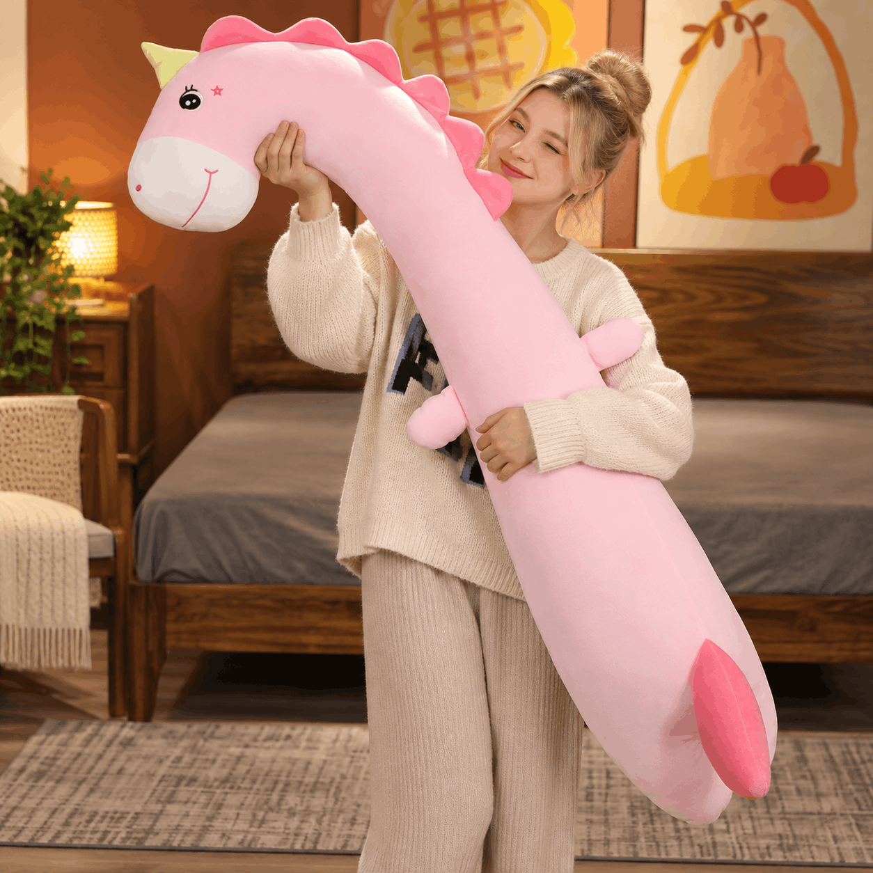 Squishmallow Fluffy Animal Body Pillow A woman in a cozy bedroom setting holds a large, long-necked pink dinosaur plush toy. The plushie has a white snout, pink horns, and a pink tail, and the woman is smiling while holding it.