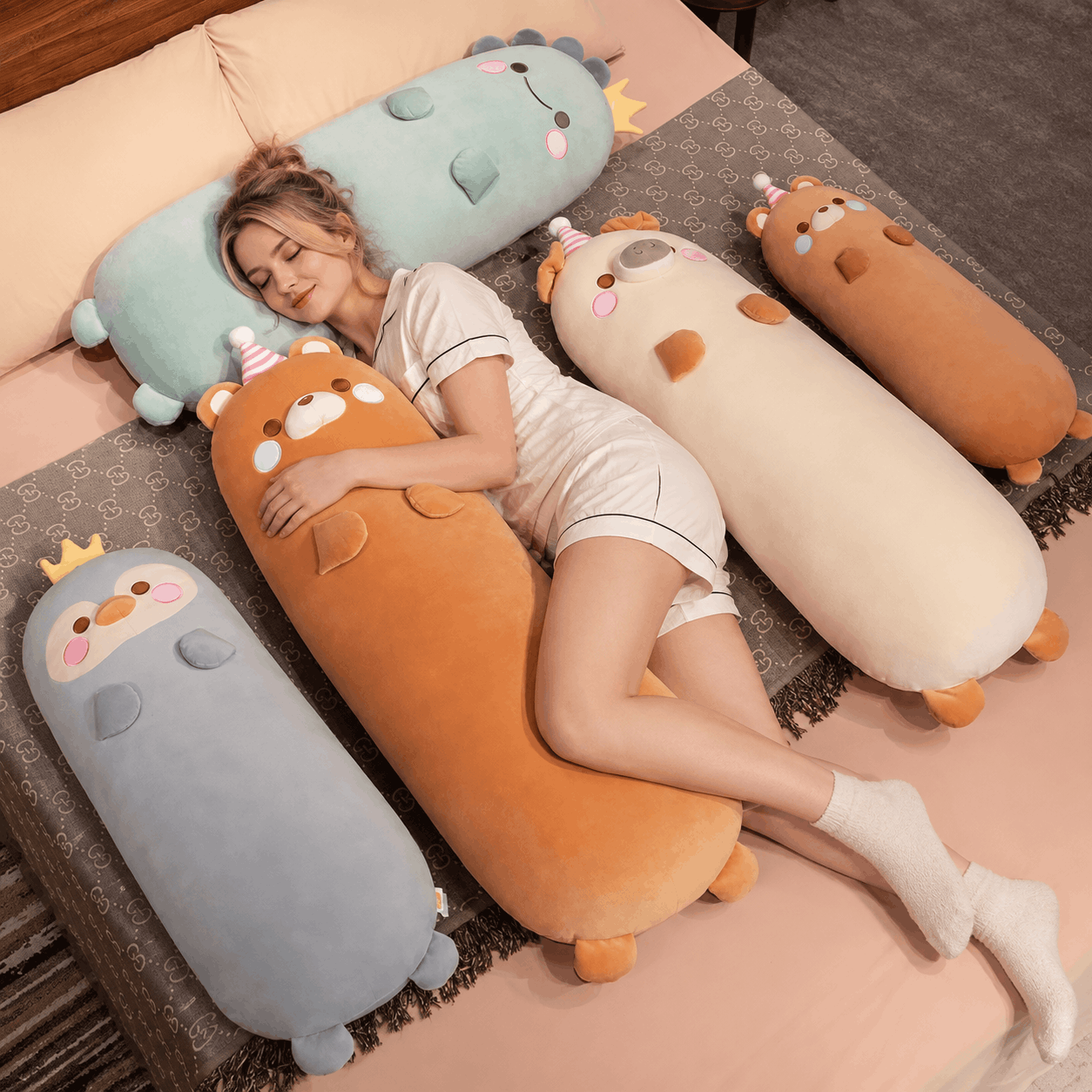 Squishmallow Long Throw Pillow Sleeper Cushion A woman lies on a bed surrounded by four large, cartoonish, animal-shaped plush pillows in pastel colors including blue, orange, cream, and gray. The pillows are decorated with cute faces, small ears, and tiny party hats.