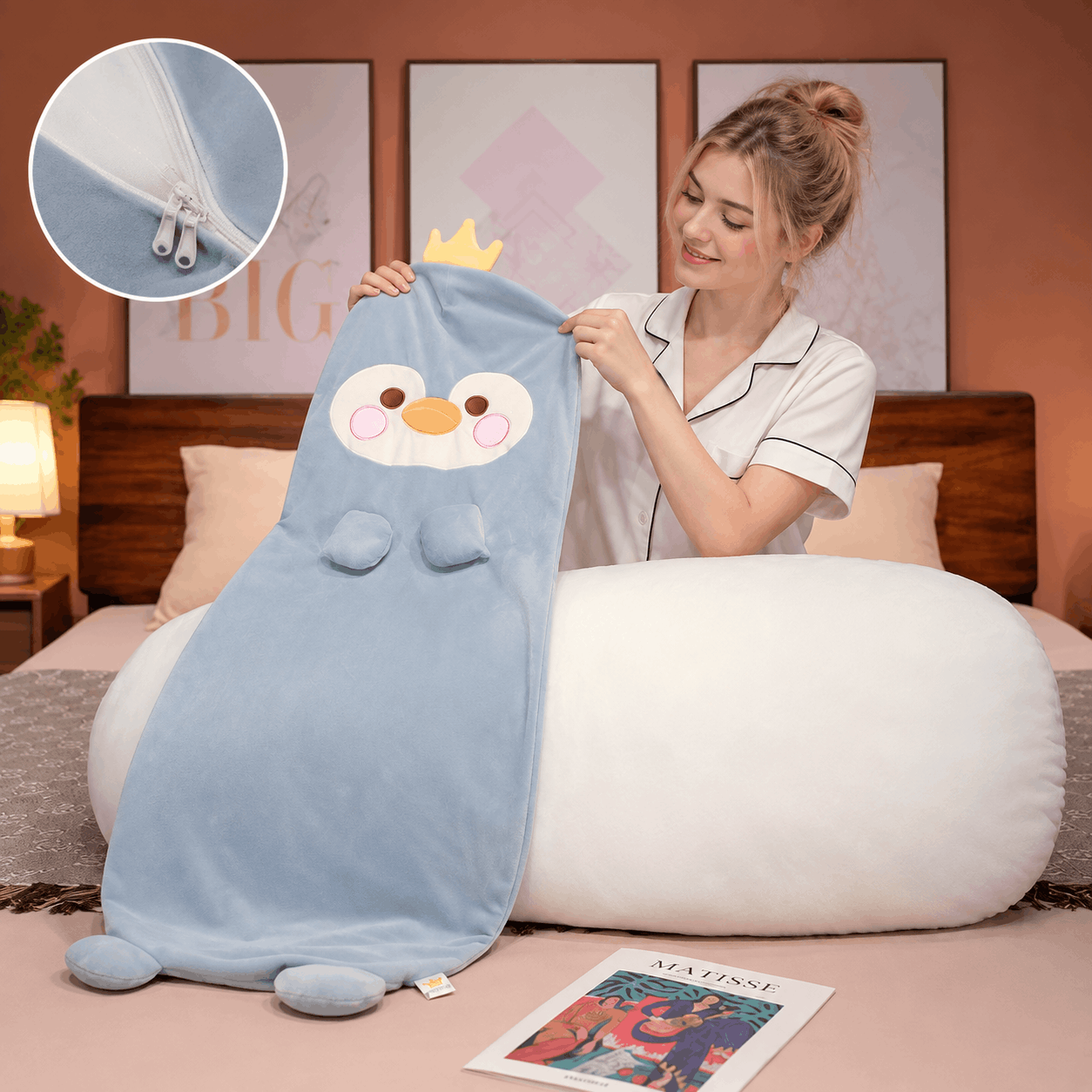 Squishmallow Long Throw Pillow Sleeper Cushion A woman in a bedroom holds a light blue plush penguin blanket with a white belly patch, pink cheeks, and a small yellow crown. The scene includes a large white oval pillow, framed wall art, a bed headboard, and a circular inset showing a close-up of the zipper detail.