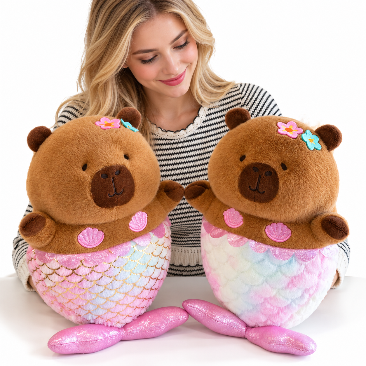 Mermaid Capybara Plush Toy – Cute Kawaii Stuffed Doll A woman with blonde hair is smiling and looking down at two plush capybara mermaids. The plush toys have brown plush bodies, colorful pastel mermaid tails in pink, blue, and white, and small flower accessories on their heads, set against a clean white background.