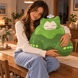 A woman sitting on a wooden dining table in a cozy, warmly lit room, holding a large green plush toy with a cartoon face and a small blue flower on its head.