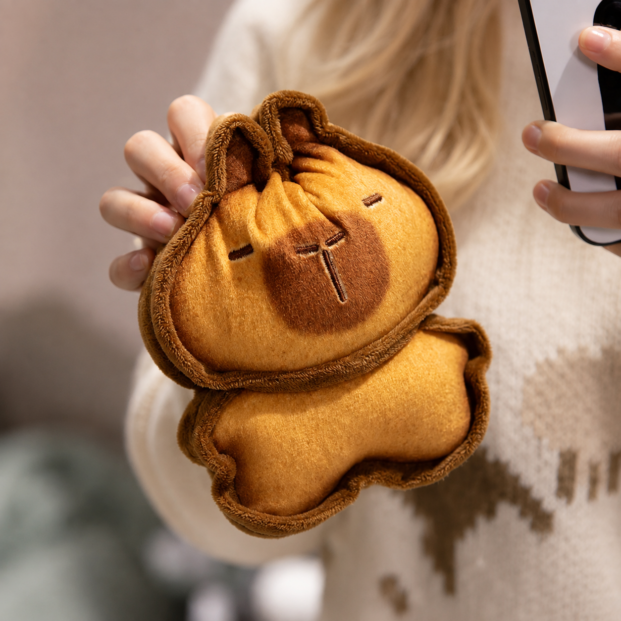 Capybara Bread Pillow – Cute Toast Plush Cushion A person holds a small plush capybara-shaped pouch or accessory in front of their face. The item is mustard yellow with a darker brown face and trim, photographed in a softly blurred indoor setting.