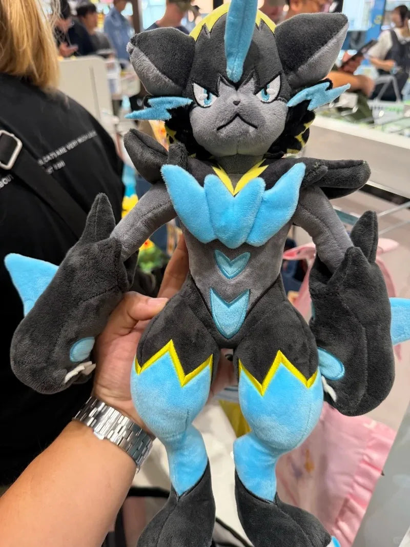 Pokemon Mega Plush Toy – Mega Lucario, Mega Greninja & Mega Starmie A dark gray and light blue plush toy with yellow accents, held up by a hand in a busy indoor setting with other people and objects blurred in the background.