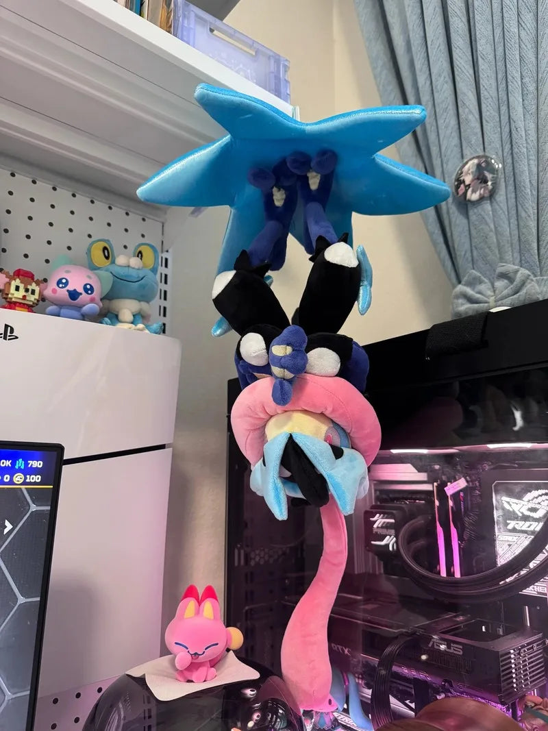 Pokemon Mega Plush Toy – Mega Lucario, Mega Greninja & Mega Starmie A colorful plush toy resembling a stylized creature with a blue star-like head, black and blue arms, and a long pink tail, displayed in a room setting next to a computer case and other small toys.