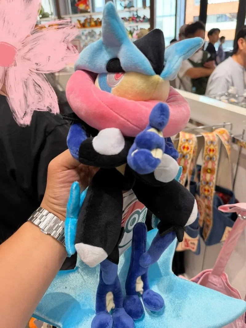 Pokemon Mega Plush Toy – Mega Lucario, Mega Greninja & Mega Starmie A plush toy of a blue and black character with a pink collar, held up by a hand in a retail store setting.