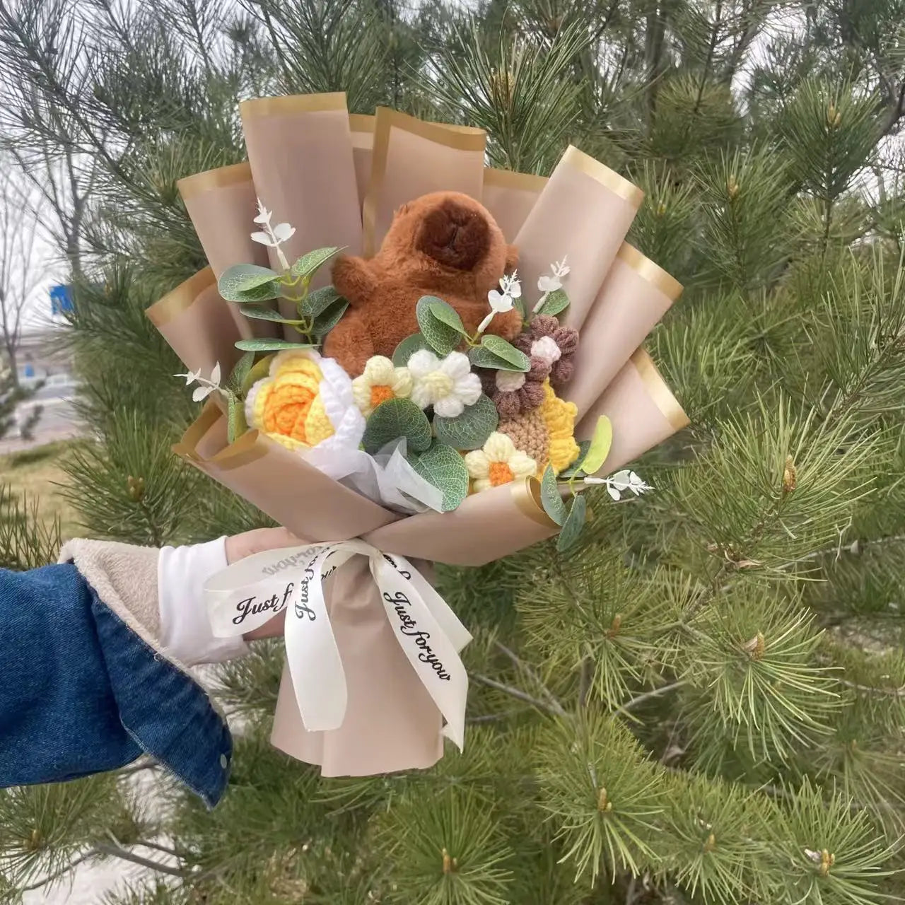 Capybara Plush with Crochet Flowers – Cute Flower Bouquet Gift Bouquet with a teddy bear and flowers held by a person against a green foliage background