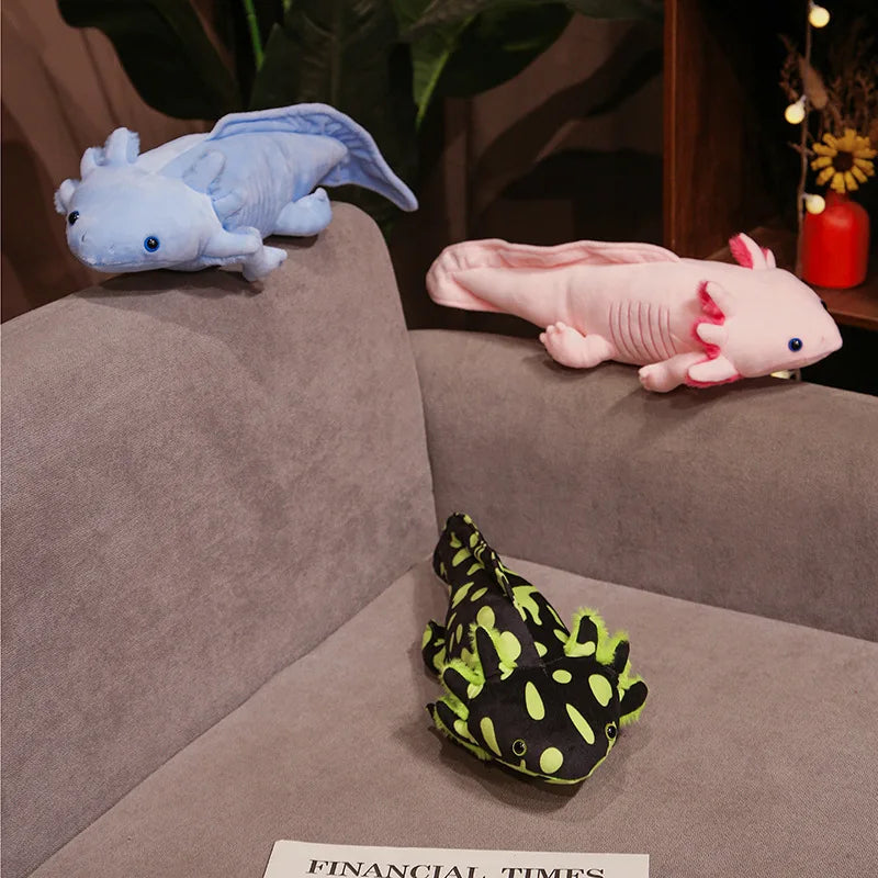 Cute Axolotl Plush Toy – Squishmallow Salamander Stuffed Animal Three plush axolotl toys in light blue, pale pink, and black with neon green spots are arranged on a gray fabric couch in a cozy indoor setting with plants and warm string lights in the background.