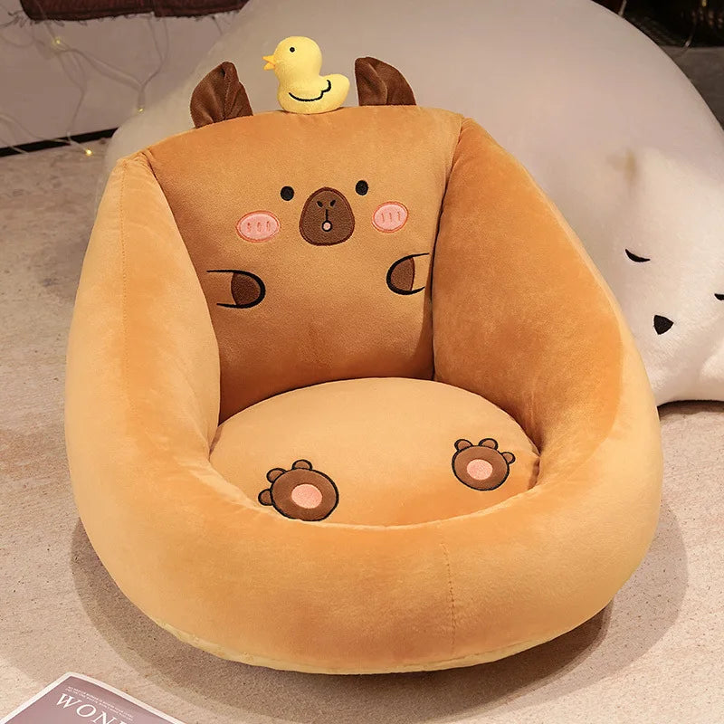 Capybara Floor Cushion – Futon Seat for Living Room & Bedroom A plush capybara-shaped chair in warm tan and cream tones, with embroidered facial features, small brown ears, a yellow duck on top, and matching foot prints on the seat cushion. The chair is photographed indoors on a light floor with a soft white plush toy in the background.