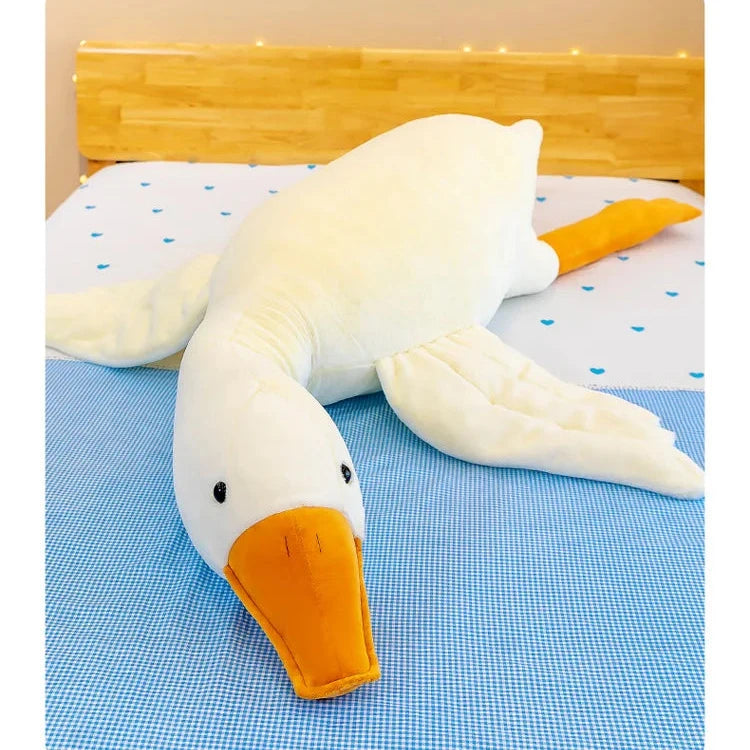 Squishmallow Plush Goose Pillow – Soft Stuffed Animal for Bed & Sofa Plush toy goose lying on a blue checkered blanket with a wooden headboard in the background.