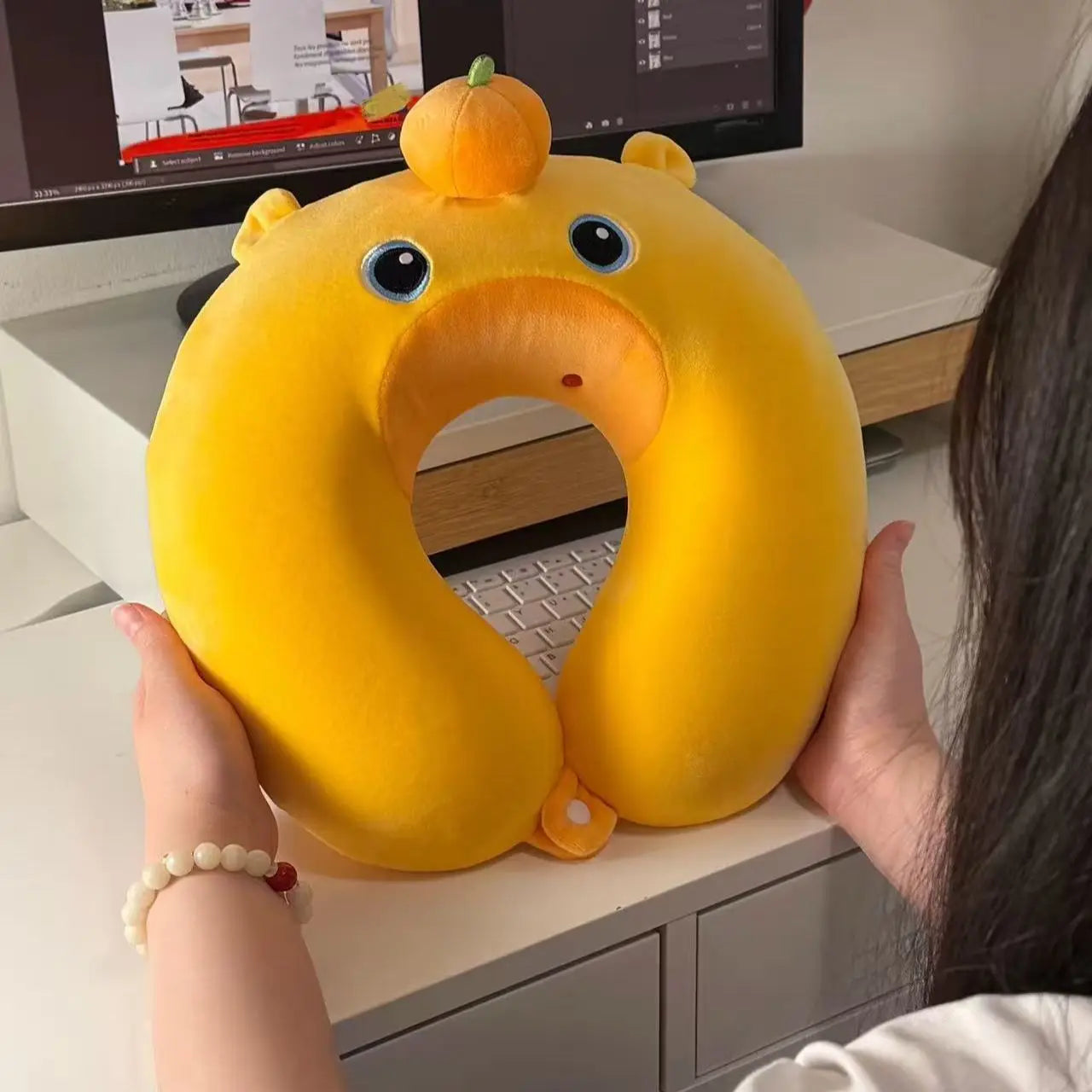 Cute Capybara U-Shaped Neck Pillow – Plush Travel Pillow A person holds a yellow duck-shaped travel neck pillow in front of a computer monitor at an office desk.
