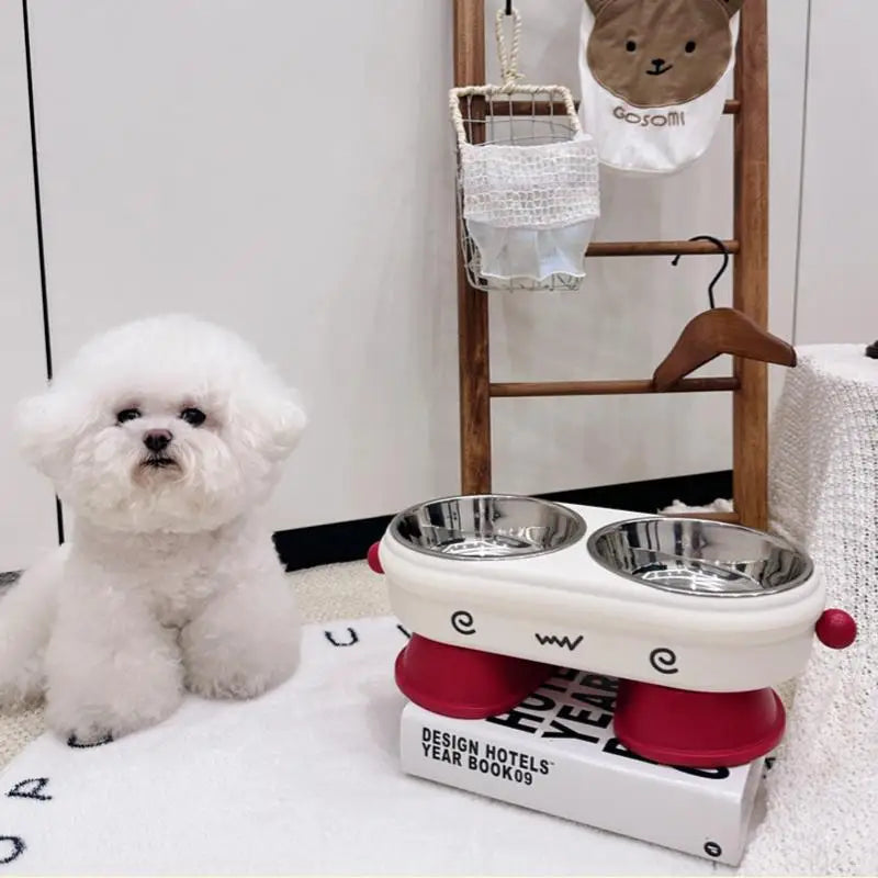 Elevated Pet Bowl Stainless Steel Anti-Slip Feeding Dish Small white dog sitting next to a double-dish pet feeder on a magazine with a ladder and baby bibs in the background.