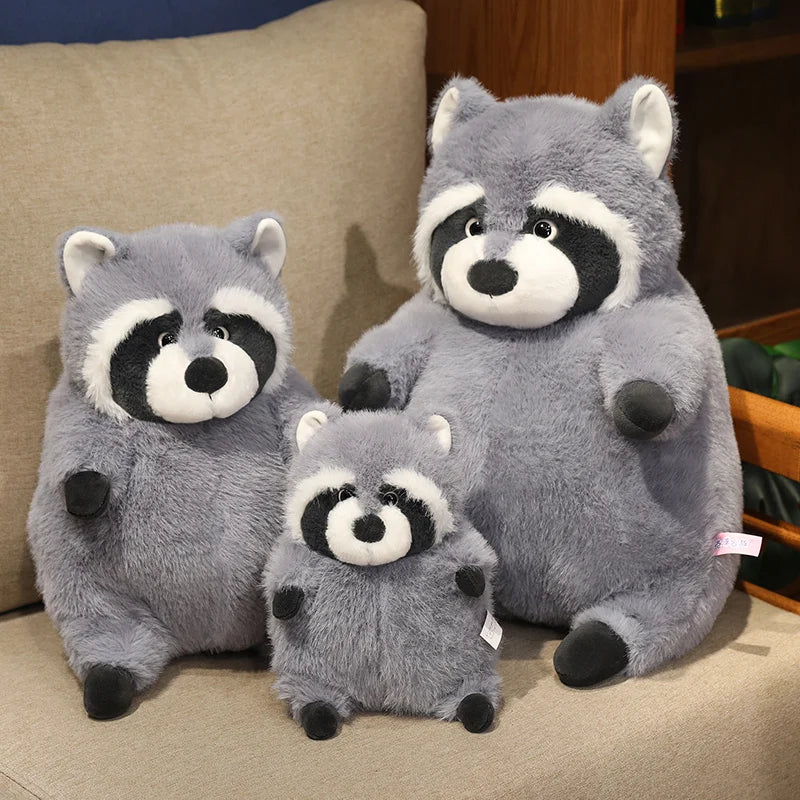 Squishmallow Grey Raccoon Plush Toy Stuffed Animal Three plush raccoons of different sizes sitting on a beige surface.
