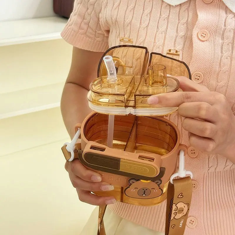 Cute Capybara Kids Water Bottle – Large Capacity Straw Cup A person in a pink cable-knit top holds a translucent amber-brown water bottle with a cartoon bear face and a matching strap. The bottle has a clear hinged lid with two straws and a compartmentalized top section.