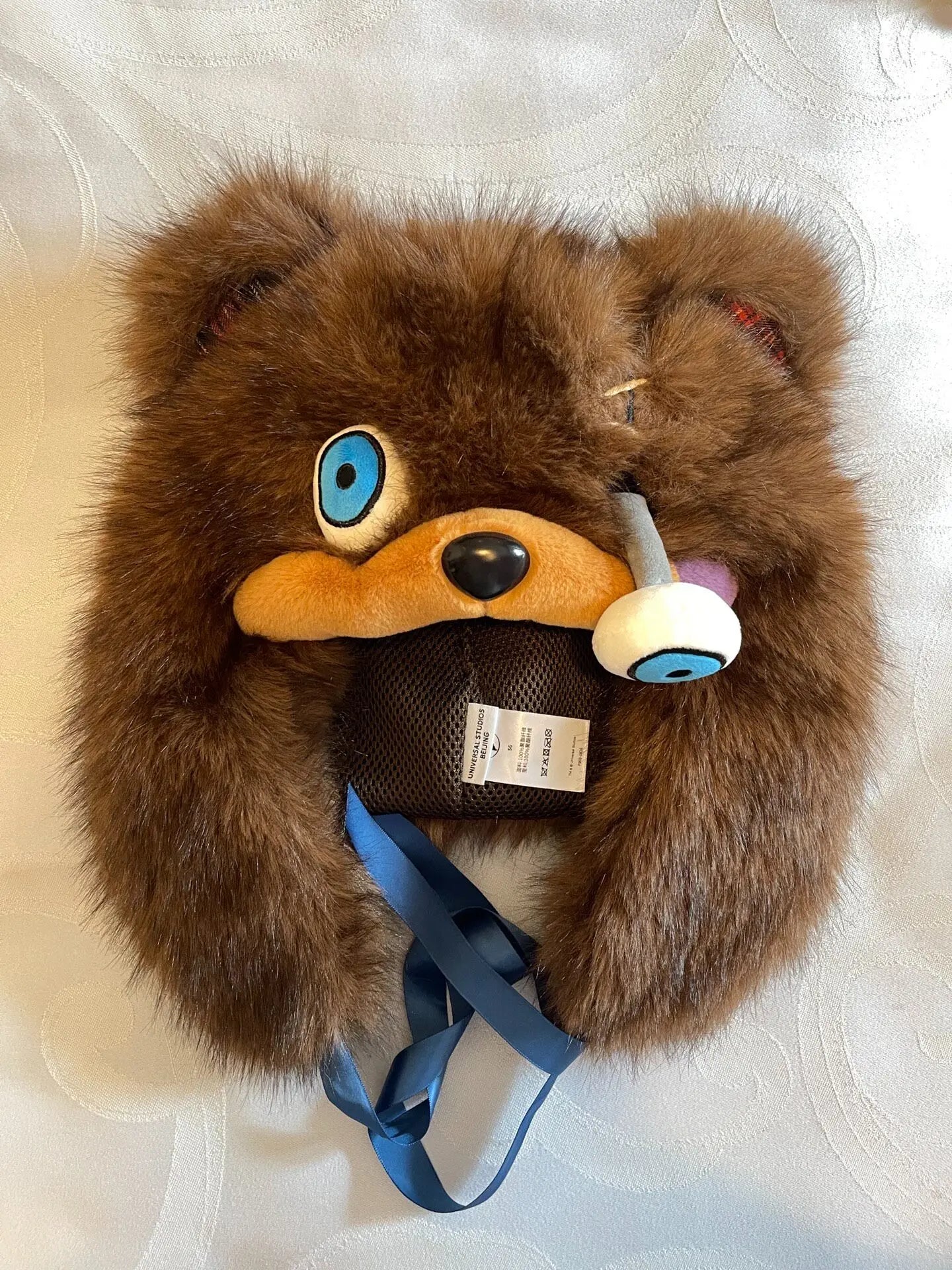 Hamikuma Bear Plush Hooded Halloween Trapper Hat A brown faux-fur teddy bear head hat or headband with blue eye details, a blue ribbon, and a small white-and-blue eye charm, laid flat on a white textured fabric background.