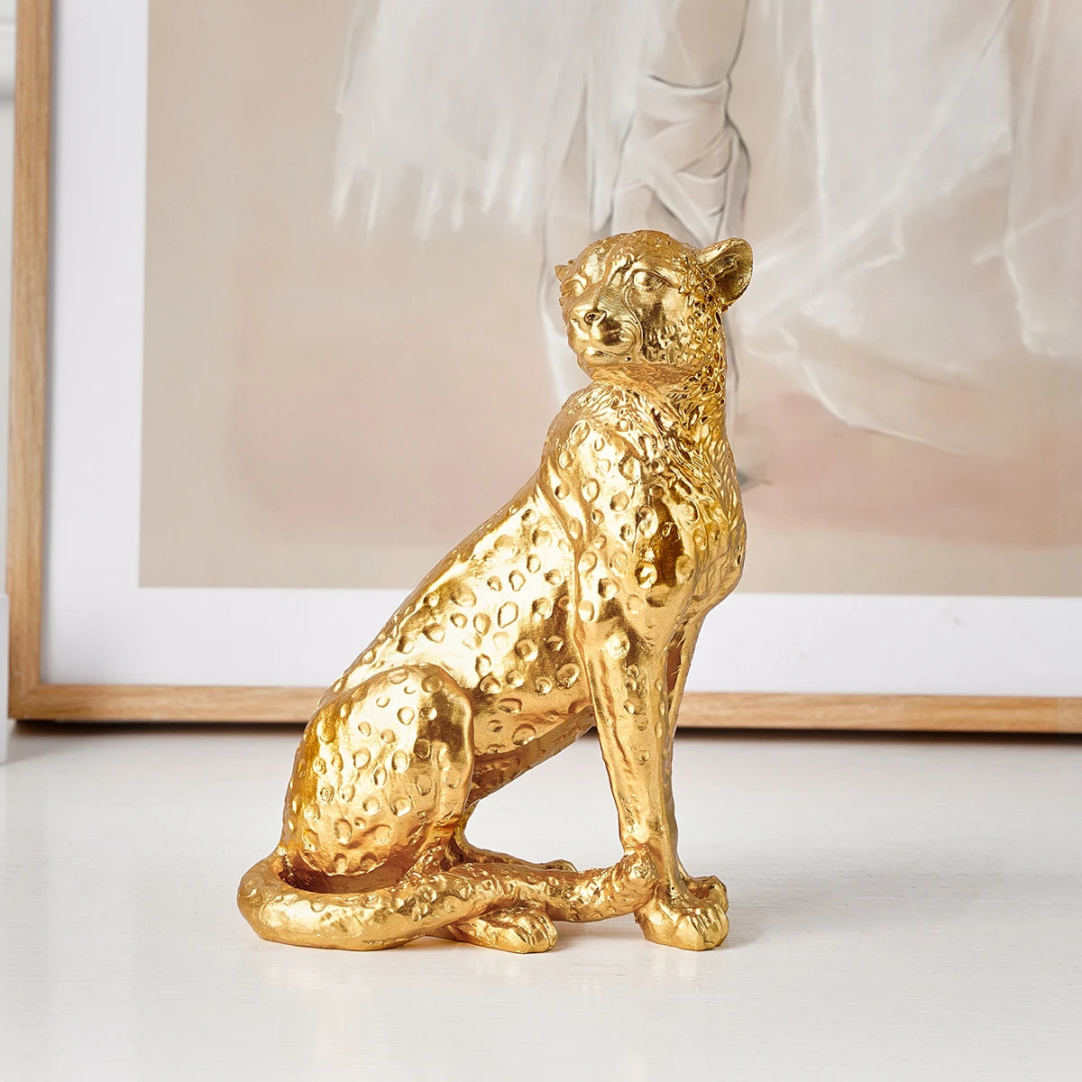Golden Leopard Figurine – Resin Animal Desktop Decor Sculpture Gold leopard figurine on a white surface with a blurred artwork in the background