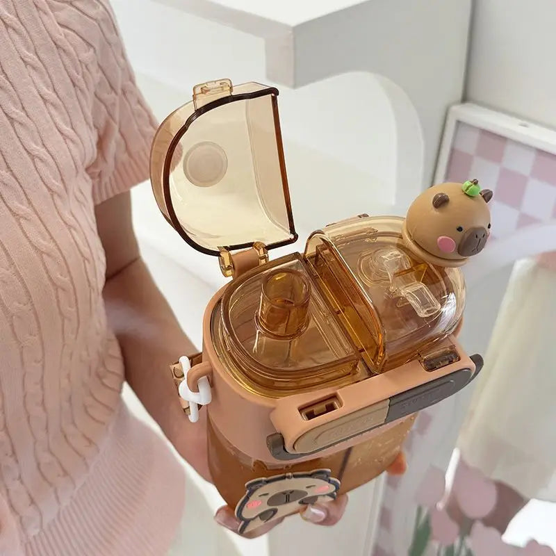 Cute Capybara Kids Water Bottle – Large Capacity Straw Cup A person in a pink knit top holds a transparent amber-colored water bottle with a cartoon cap and a small animal character on top, shown in a bright indoor setting.