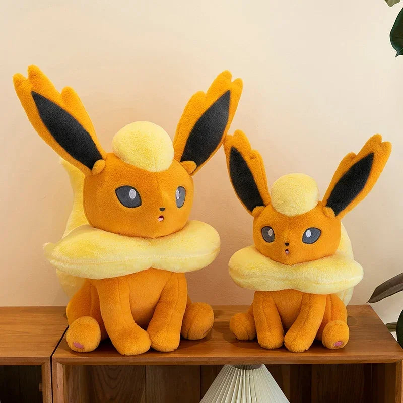 Pokemon Eevee Plush Toy – Vaporeon, Flareon & Jolteon Soft Stuffed Animal Two yellow and orange plush toy characters with black-tipped ears and cream collars, sitting on a wooden shelf against a beige wall.