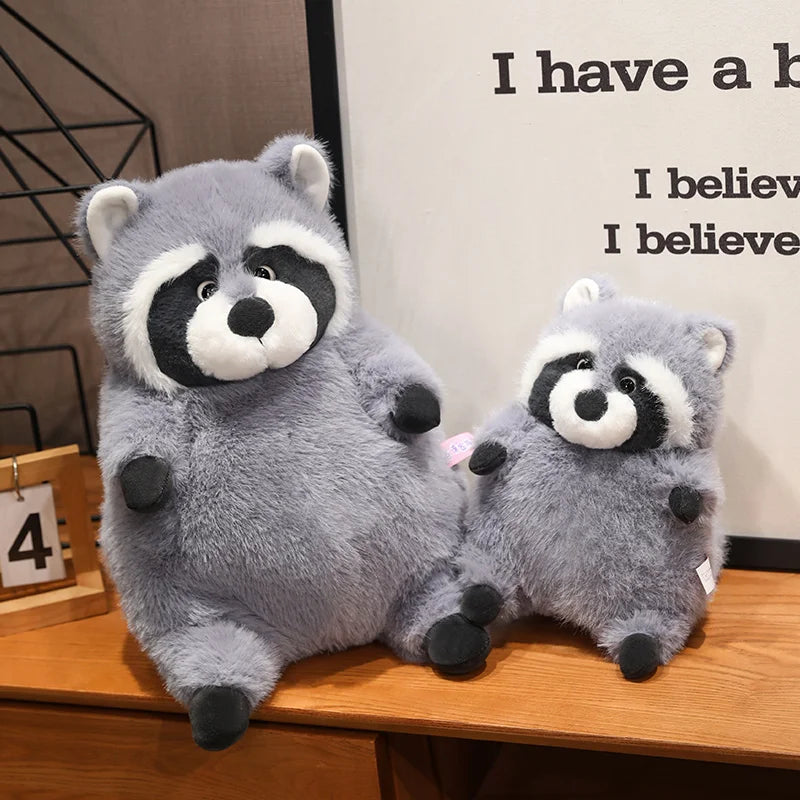 Squishmallow Grey Raccoon Plush Toy Stuffed Animal Two gray raccoon plush toys on a wooden surface with a neutral background.
