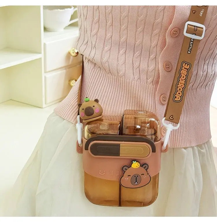 Cute Capybara Kids Water Bottle – Large Capacity Straw Cup A pink and brown bear-themed crossbody water bottle bag worn by a person in a pink cable-knit cardigan and white skirt. The bag features a cute cartoon bear face, a small plush bear accessory, and a brown adjustable strap with a white buckle.