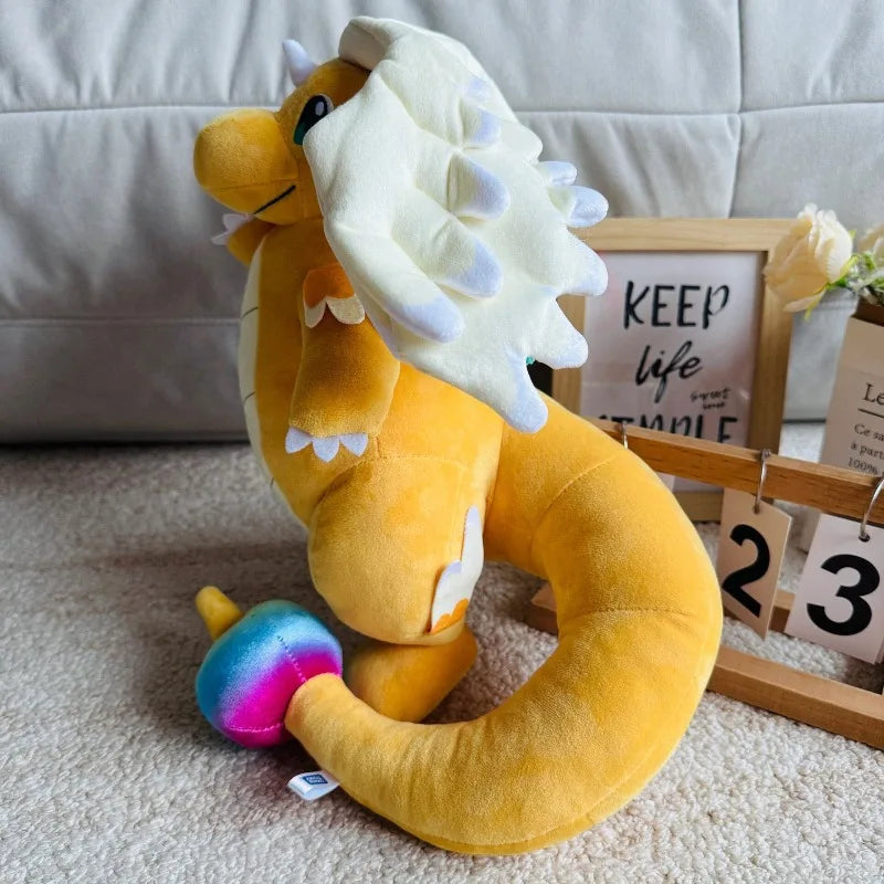 Pokemon Dragonite Plush Toy – Large Soft Stuffed Doll A yellow plush toy dragon with a white horned crest sits on a beige carpet in front of a light gray sofa. A small colorful mushroom-shaped accessory is placed near its tail, with a framed sign and wooden calendar blocks in the background.