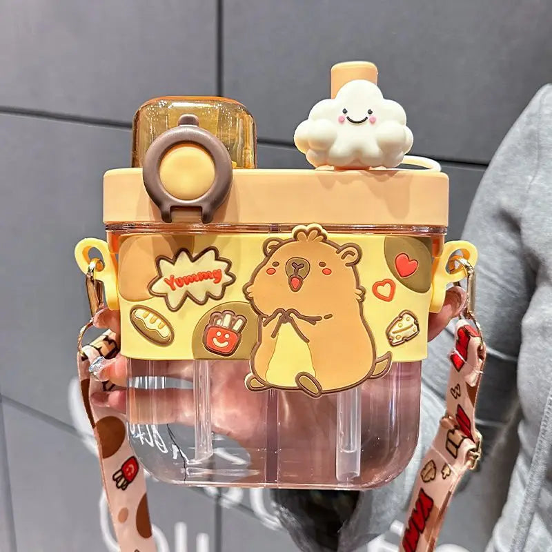 Capybara Kids Water Bottle – Cartoon Straw Cup A clear plastic water bottle with a yellow cartoon animal and food-themed front cover, a beige lid, a small white cloud topper, and a patterned shoulder strap, being held by a person against a gray tiled background.