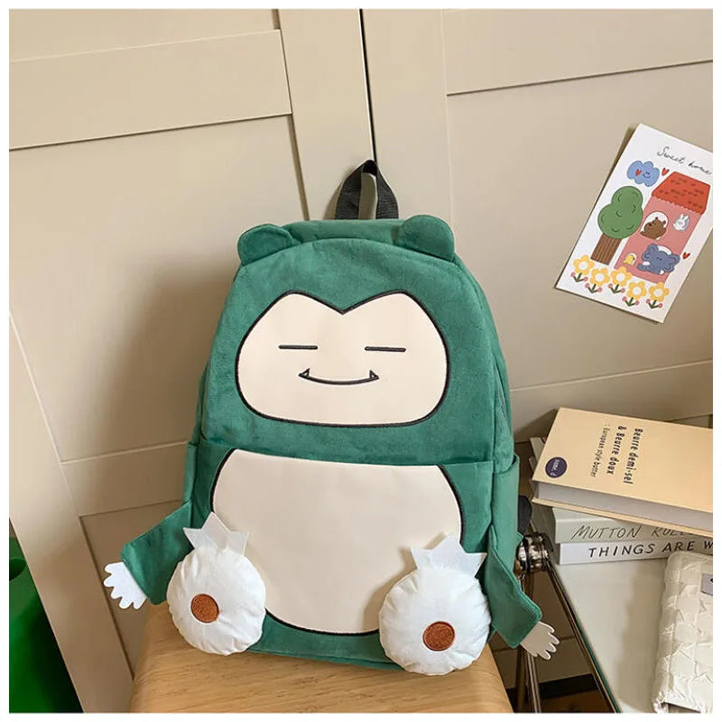 Snorlax Pokemon Backpack Large Capacity Casual Bag A green plush backpack shaped like a cartoon character with a large cream face and two small white plush feet, displayed on a chair in a room setting next to books and a card.