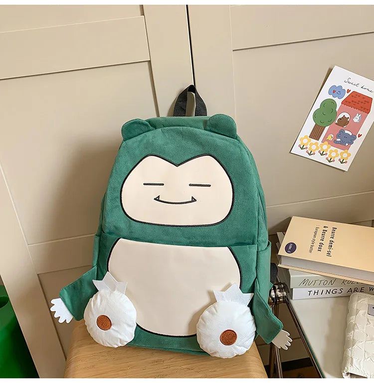 Snorlax Pokemon Backpack Large Capacity Casual Bag A plush green and white character backpack shaped like a cartoon creature, displayed on a wooden surface in front of a beige paneled wall with books and a small card nearby.