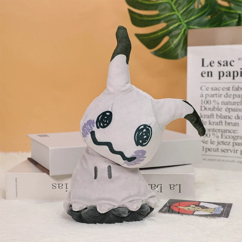 Pokemon Mimikyu Plush – Shiny Stuffed Toy A plush toy of a ghost-like character with a white body, dark gray pointed ears and feet, and a stitched smiling face, styled on books with a book leaf in the background.