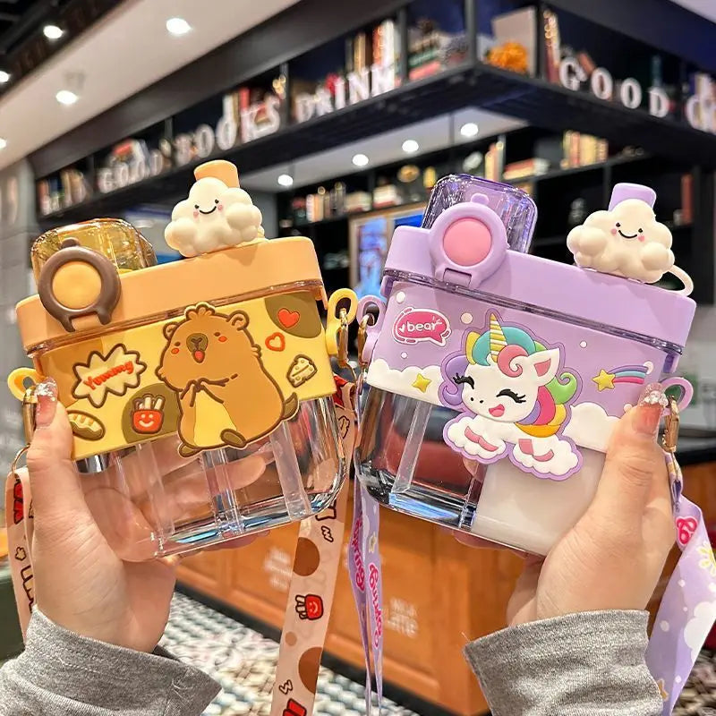 Capybara Kids Water Bottle – Cartoon Straw Cup Two cute, transparent plastic water bottles with colorful silicone covers are held up by two hands in a cafe setting. The bottle on the left is yellow with a capybara and cartoon sticker design, and the bottle on the right is lavender with a unicorn and rainbow design.