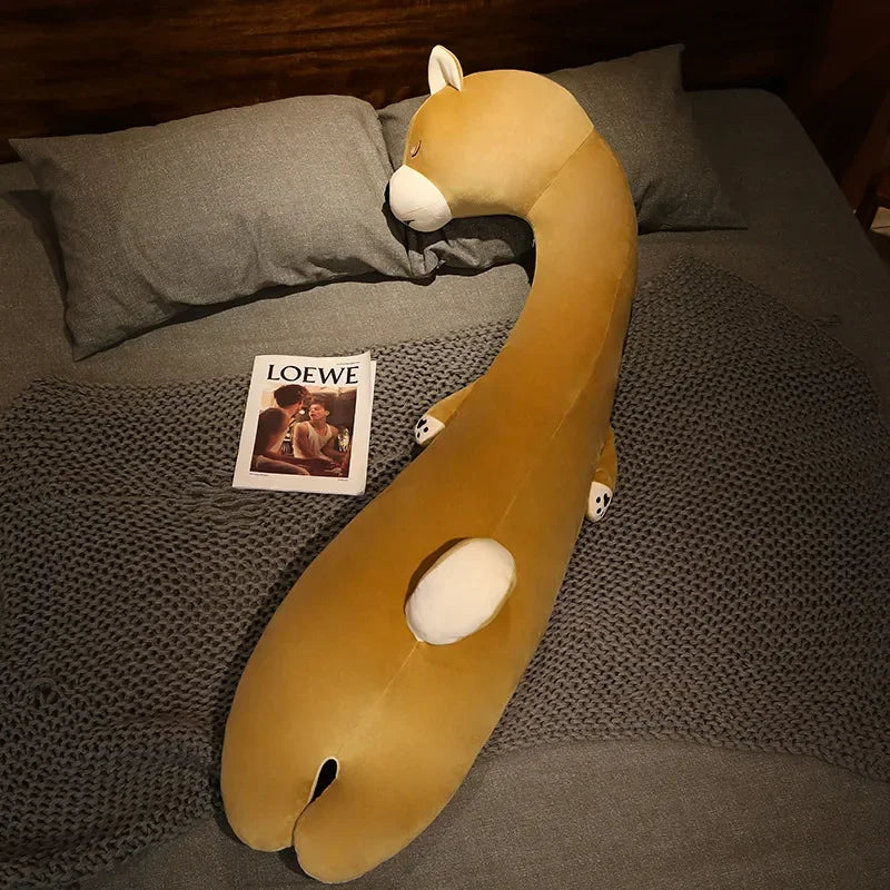 Squishmallow Fluffy Animal Body Pillow A long, curved mustard-yellow plush animal toy with white accents lies on a gray bed with pillows and a knit blanket. A magazine titled LOEWE is placed beside it.