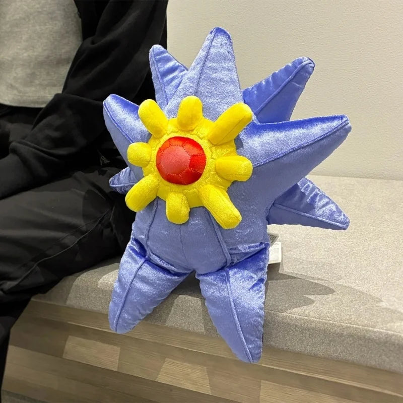 Pokemon Mega Plush Toy – Mega Lucario, Mega Greninja & Mega Starmie A plush star-shaped pillow in shades of blue, yellow, and red, styled on a gray bench next to a person wearing a black jacket.