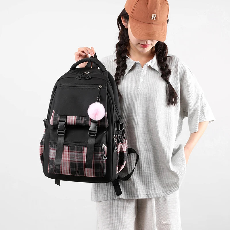 Capybara Bubble Tea Backpack – School Bag for Teens A person wearing a light gray oversized polo shirt and matching sweatpants holds up a black backpack with pink plaid accents and a fluffy pink pom-pom charm. The person is also wearing a brown baseball cap with a white 'R' logo, set against a plain light gray studio background.