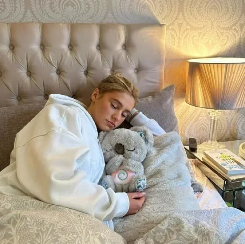 Baby Breathing Otter Plush with Light & Sound Person sleeping in bed with a teddy bear, wearing a robe, in a cozy bedroom setting.
