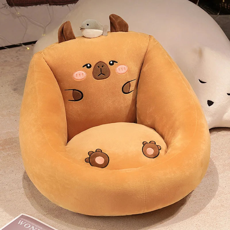 Capybara Floor Cushion – Futon Seat for Living Room & Bedroom A plush capybara-shaped chair in warm camel-brown velvet, featuring embroidered facial details, small brown ears, and a small plush bird on top, photographed indoors on a light floor with a white plush toy partially visible behind it.