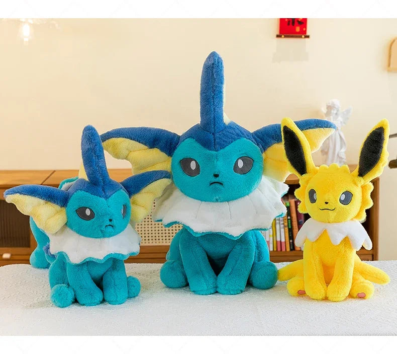 Pokemon Eevee Plush Toy – Vaporeon, Flareon & Jolteon Soft Stuffed Animal Three plush toys arranged on a white surface in an indoor room: two blue-and-yellow Eeveo plushies and one yellow Eevee plushie, with a blurred bookshelf and wall in the background.