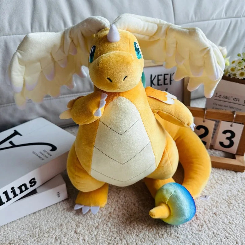 Pokemon Dragonite Plush Toy – Large Soft Stuffed Doll A yellow and cream-colored plush dragon toy with large wings, blue eyes, and a small iridescent mushroom on its tail, posed on a beige carpet in a styled indoor setting with books and a calendar.
