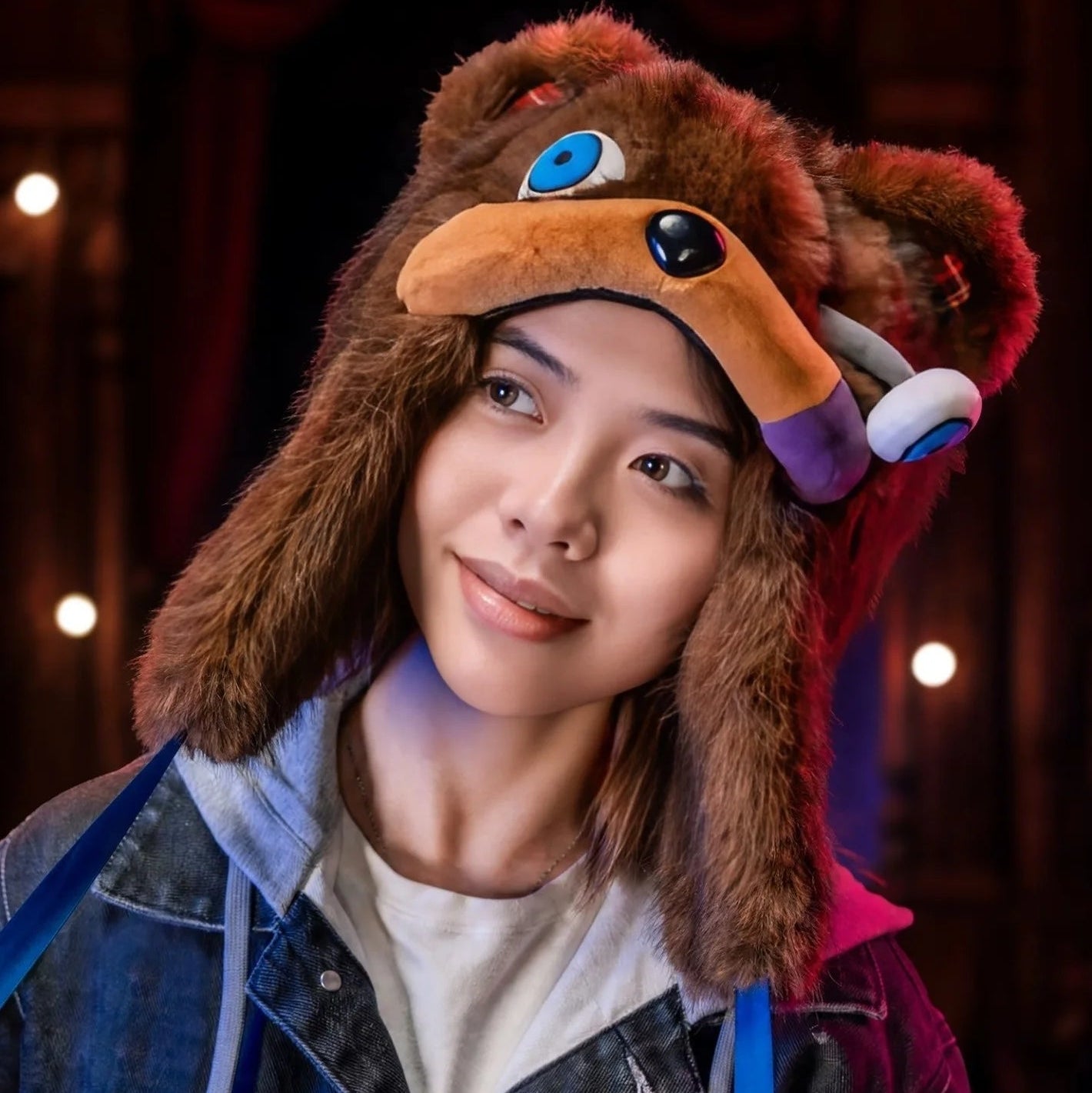Hamikuma Bear Plush Hooded Halloween Trapper Hat A person wearing a brown furry animal-themed hood with a plush face, blue eyes, and a purple ear, photographed in a dim indoor setting with warm lights.