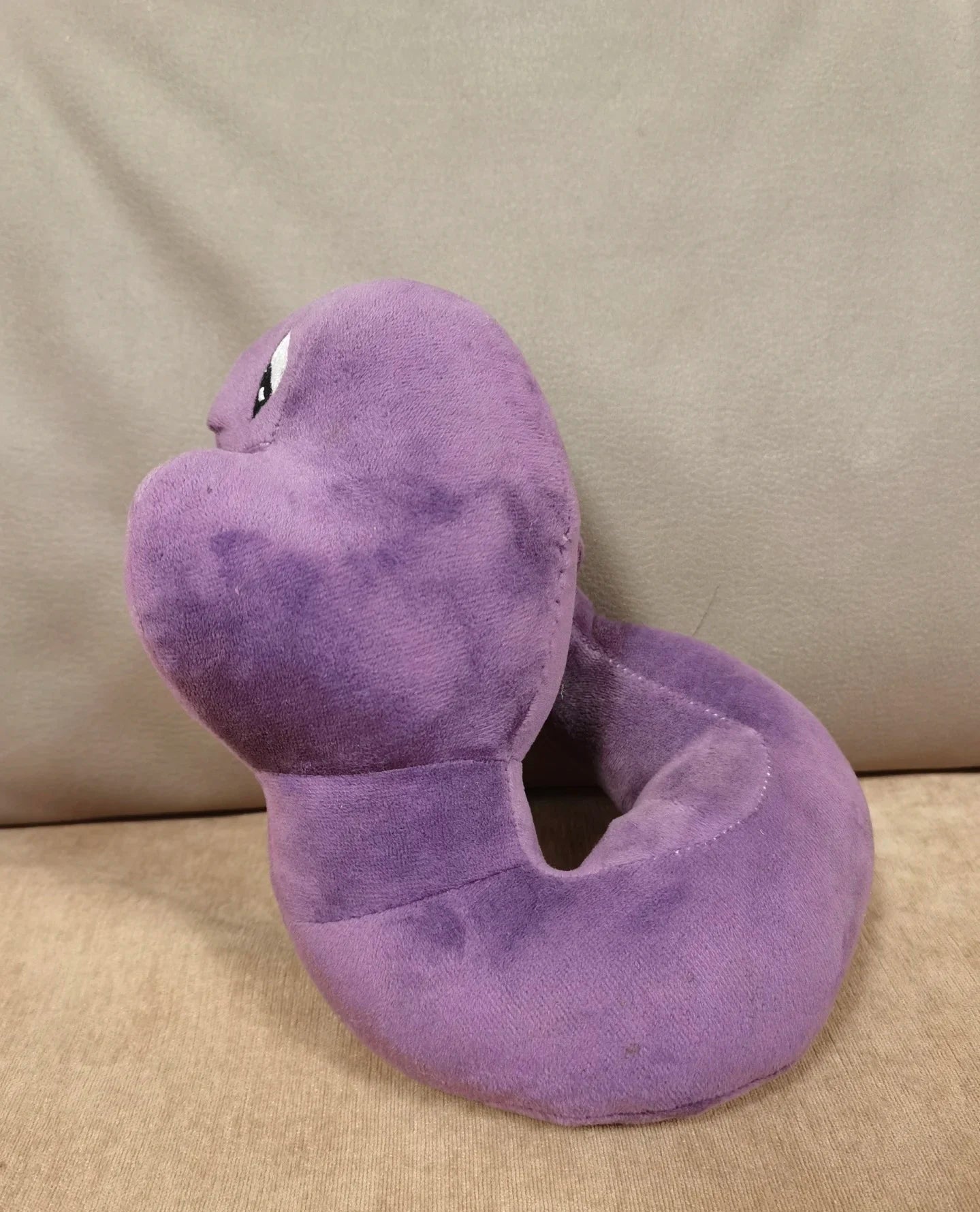 Pokemon Arbok Plush Toy – Ekans Evolution Stuffed Animal A purple plush snake-shaped neck pillow with embroidered eyes, resting on a beige fabric surface against a matching beige background.
