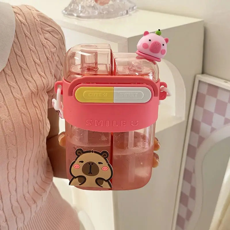 Cute Capybara Kids Water Bottle – Large Capacity Straw Cup A person holds a pink double-straw water bottle with a capybara sticker and a small pig-shaped straw topper in a bright indoor setting.