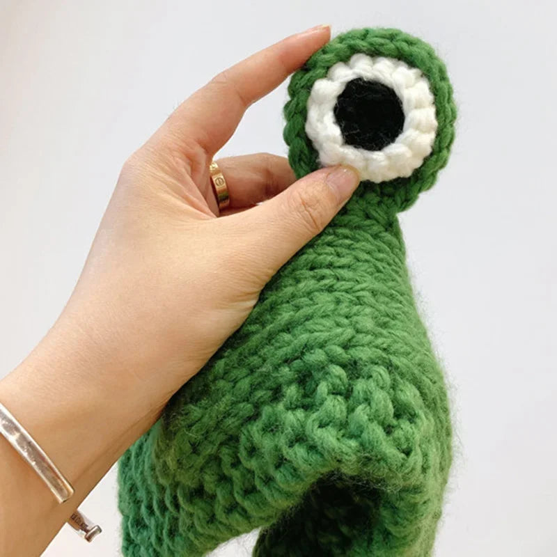 Cute Green Frog Crochet Beanie Hat – Knitted Winter Costume Cap Hand holding a green knitted toy with a white circle and black center on a light gray background