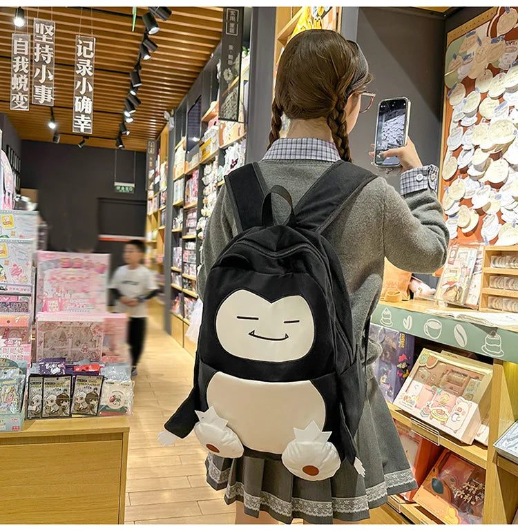 Snorlax Pokemon Backpack Large Capacity Casual Bag A person wearing a gray blazer and pleated skirt stands in a retail store, viewed from behind, wearing a black penguin backpack with a large white belly patch and small white feet details.