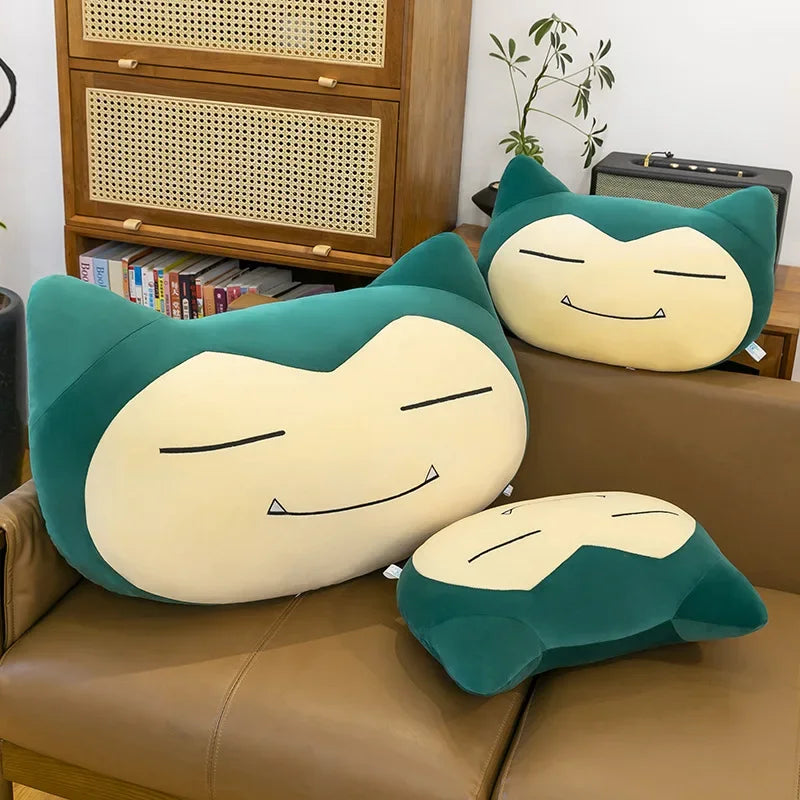 Pokemon Snorlax Plush Pillow – Cute Oversized Stuffed Animal Three Snorlax plush toys on a brown couch with a wooden cabinet and books in the background.