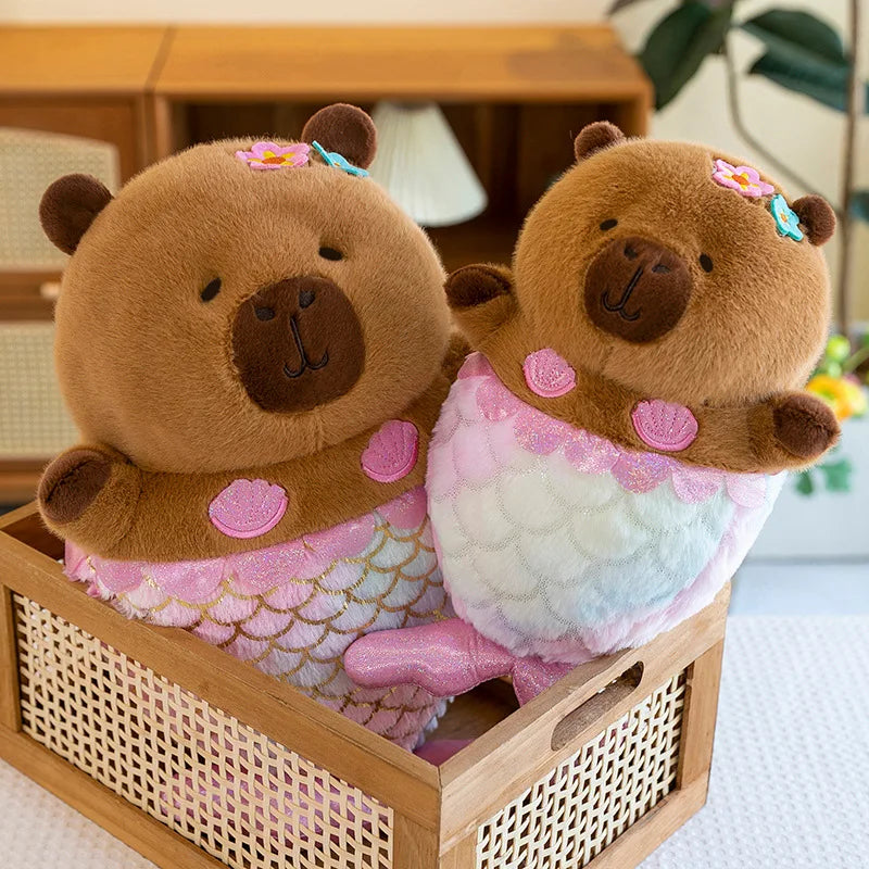 Mermaid Capybara Plush Toy – Cute Kawaii Stuffed Doll Two plush capybara toys sitting in a wooden rattan box. One capybara wears a pink and gold mermaid tail, and the other wears a pink and white mermaid tail with a pastel gradient.